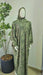 Green patterned prayer dress on a mannequin against a plain wall with a plant in the background